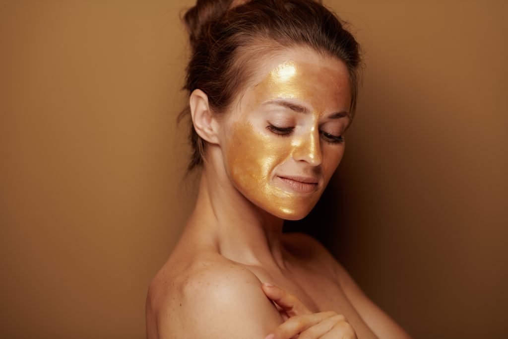 Portrait of modern middle age woman with golden cosmetic face mask on bronze background.